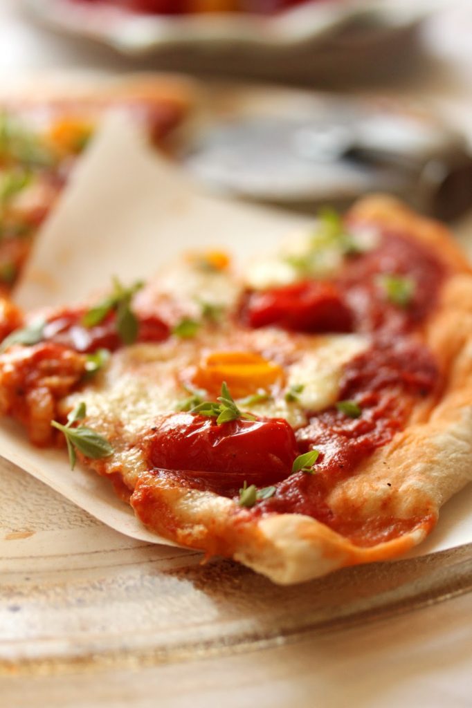 Part de pizza posé sur une feuille de papier cuisson et garnie de coulis de tomates, mozzarella et tomates cocktail rouges et jaunes coupées en deux