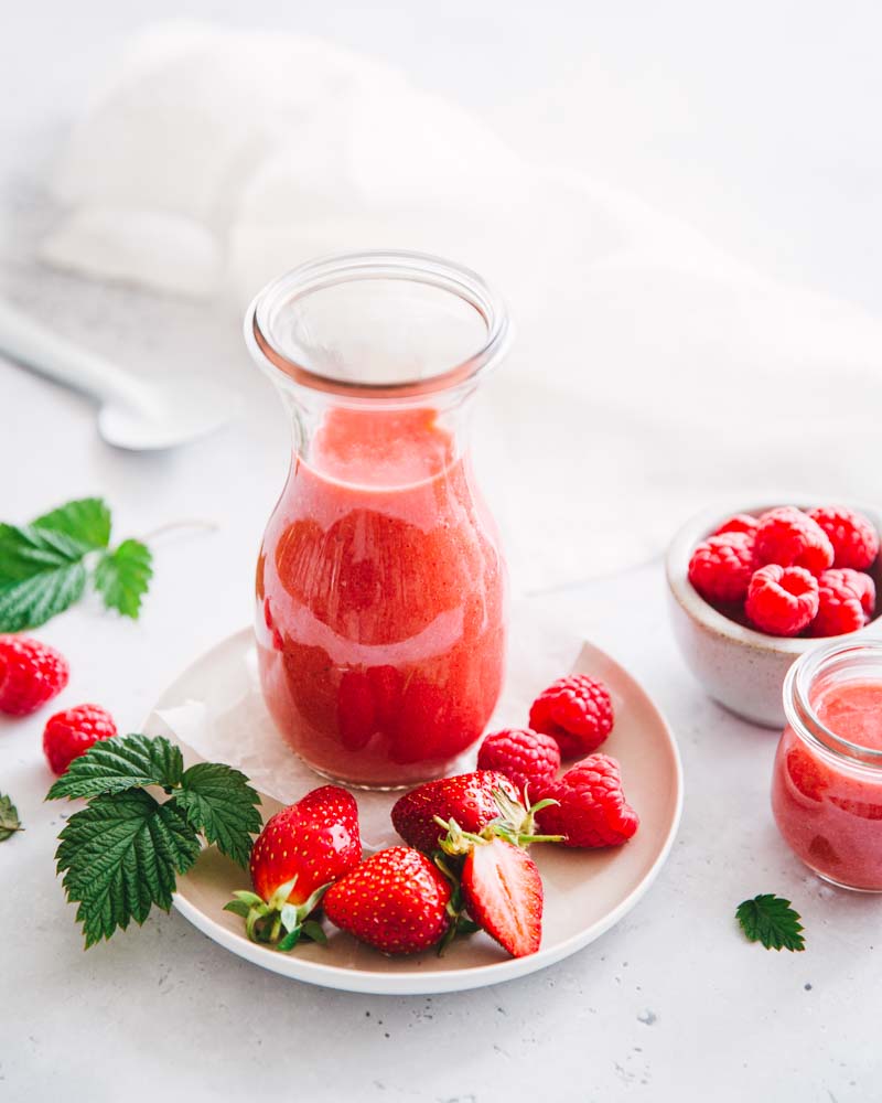 Petite bouteille en verre, posée sur une assiette avec des fruits frais, contenant un smoothie frais aux fraises et framboises fait maison. 