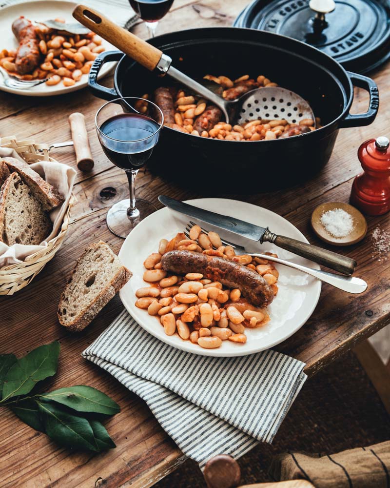 Assiettes garnies de haricots tarbais à la sauce tomate cuisinés en cocotte avec des saucisses, sur une table en bois. Ambiance rustique. 
