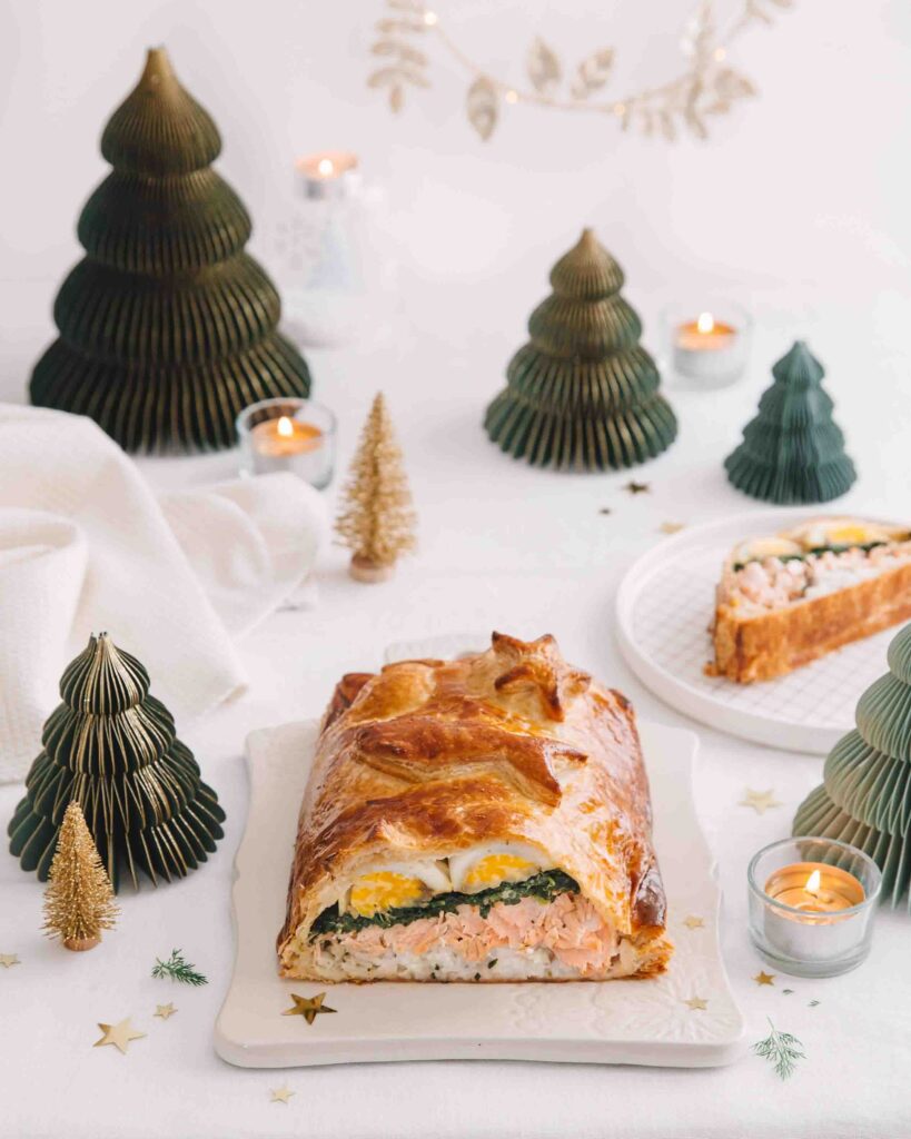 Koulibiac de saumon tranché et posé sur une planche en porcelaine dans un décor festif avec des sapins de noël. 