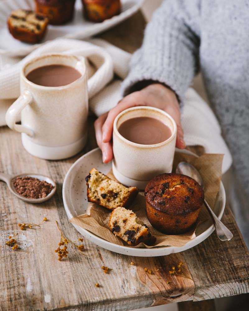 Main saisissant un tasse de chocolat chaud végétal avec une assiette de muffins ig bas aux pépites de chocolat pour le goûter. 