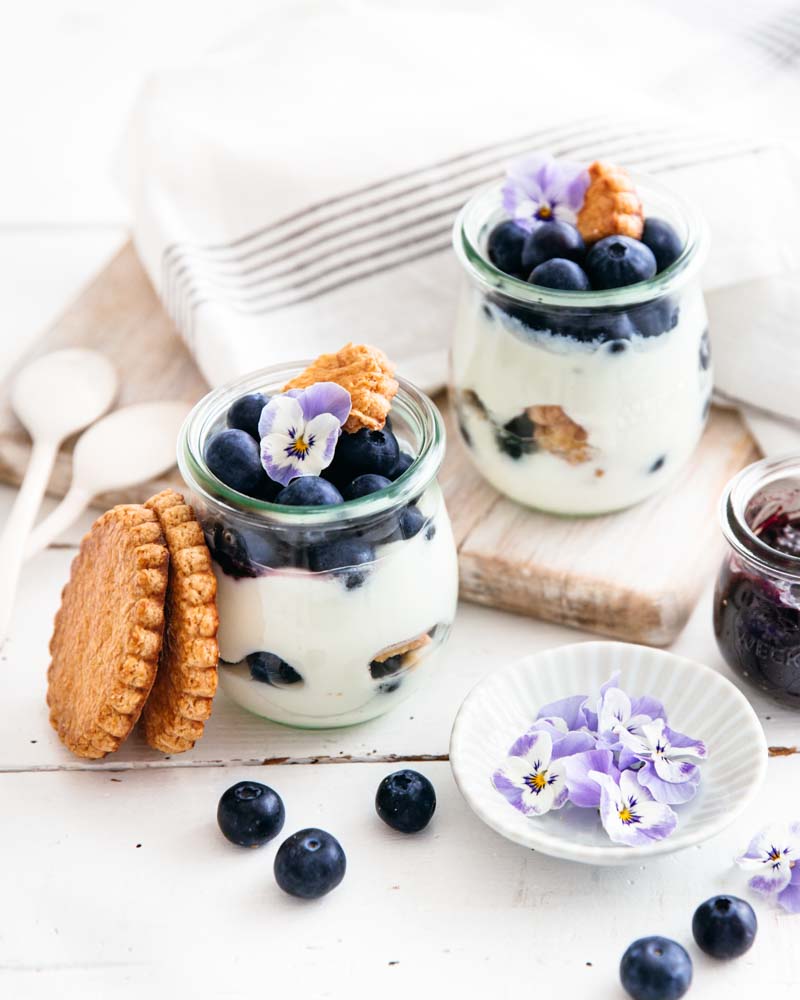 Dessert gourmand dans des pots en verre composés de yaourt, confiture, myrtilles et biscuits secs dans une ambiance épurée