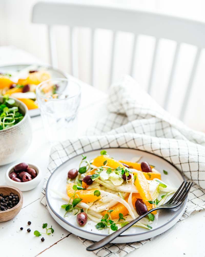 Assiette garnie d'une salade de fenouil, orange et olives noires dans une ambiance lumineuse avec une table rustique en bois blanc. 