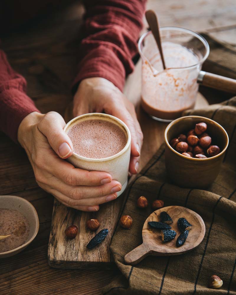 Mains autour d'un mug de latte praliné dans une ambiance chaleureuse. Une recette saine à base de noisettes, cacao cru et épices. 