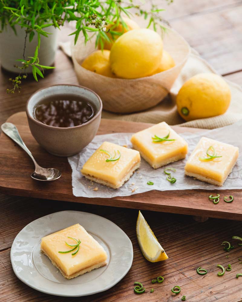 Carrés au citron présentés sur une planche en bois accompagnés d'une tasse de thé et de citron dans le fond de la photo. Ambiance cosy et douce pour un goûter gourmand. 