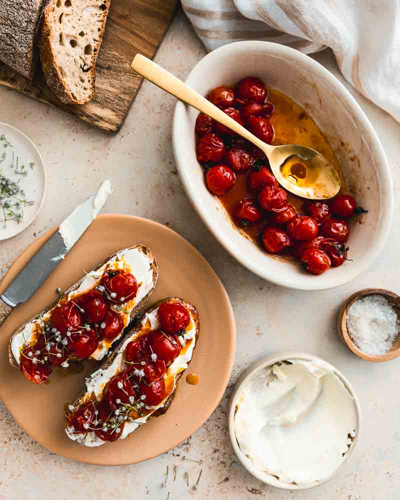 Vue de haut d'une assiette garnie de deux tartines de pain recouvertes de fromage frais, tomates rôties et fleur de poireau. A côté se trouvent les ingrédients qui composent cette recette. 