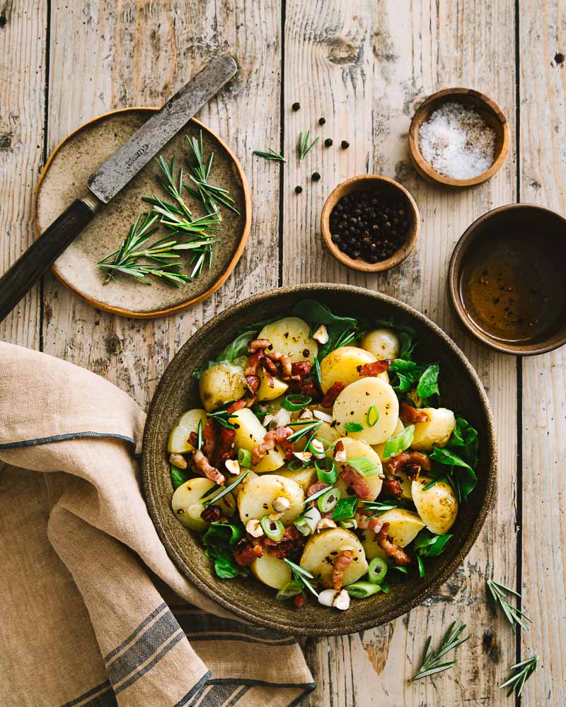Ambiance rustique vue de haut d'une assiette de salade de pommes de terre primeur aux lardons, oignons nouveaux, noisettes et salade verte. Accompagnée des ingrédients de la sauce salade qui va avec. 