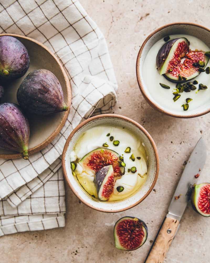 Photo vue de haut de deux bols de dessert aux figues et pistaches dont un garni de miel. Une assiette de figues à côté