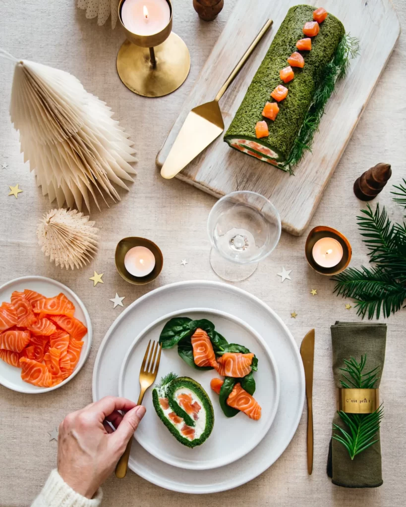 vue de haut d'une bûche salée pour noël : génoise aux épinards et garniture au fromage frais, aneth et saumon fumé. Dans l'assiette, une tranche est servie et accompagnée de lamelles de saumon et de pousses d'épinards