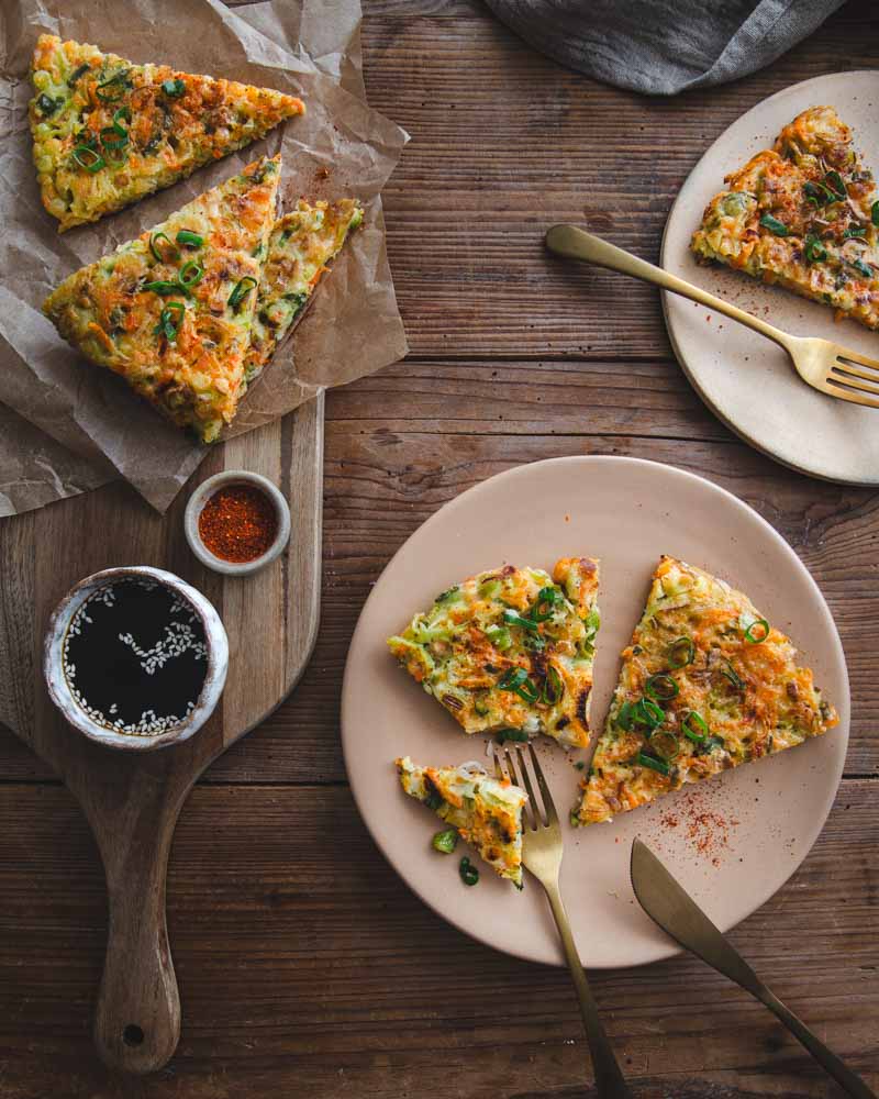Galettes coréenne aux légumes d'hiver - poireaux et carottes - posés sur des assiettes en céramique beige. A gauche, une planche en bois avec un papier cuisson froissé et des parts de galette, une coupelle de sauce soja et de Piment d'Espelette. Ambiance rustique avec table en bois. 