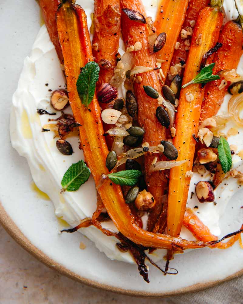 Gros plan sur des carottes rôties au four additionnées d'un granola salé aux noisettes et aux graines de courge. Sous les légumes, une épaisse couche de yaourt à la grecque onctueux. 