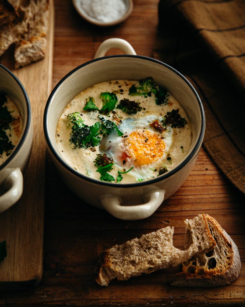 Oeuf cocotte Air Fryer aux brocolis et au persil avec de la crème de soja dans un ramequin en céramique beige avec un liseré noir et des petites anses. 