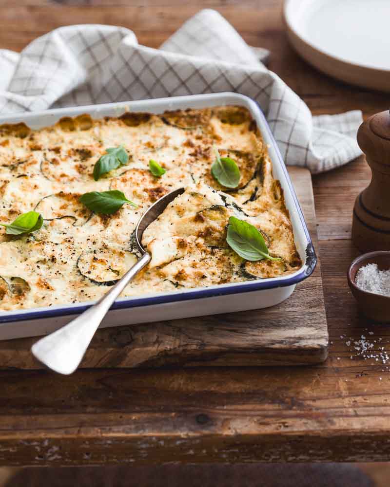 Gratin de courgettes végétal cuit dans un plat Falcon à liseré bleu, posé sur une planche en bois, dans une ambiance rustique. Prêt pour la dégustation avec une cuillère dans le plat. 