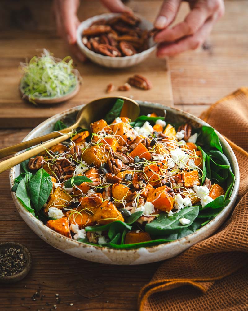 Main dans le fond de la photo qui va déposer des noix de pécan dans le saladier rempli de légumes rôtis à l'air Fryer, de salade d'épinards, de féta, de graines germées et de courge. 