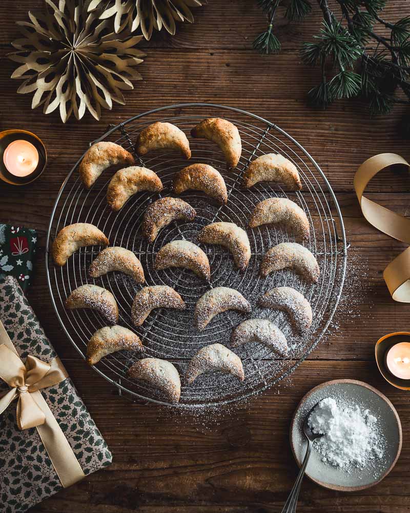 Vue de haut de sablés de noël aux amandes sans gluten comme des Vanille Kipferln dont certains sont saupoudrés de sucre glace. Aujourd'hui des bougies, des paquets cadeaux, une coupelle de sucre glace, un ruban doré, des rosaces en papier doré et un peu de sapin