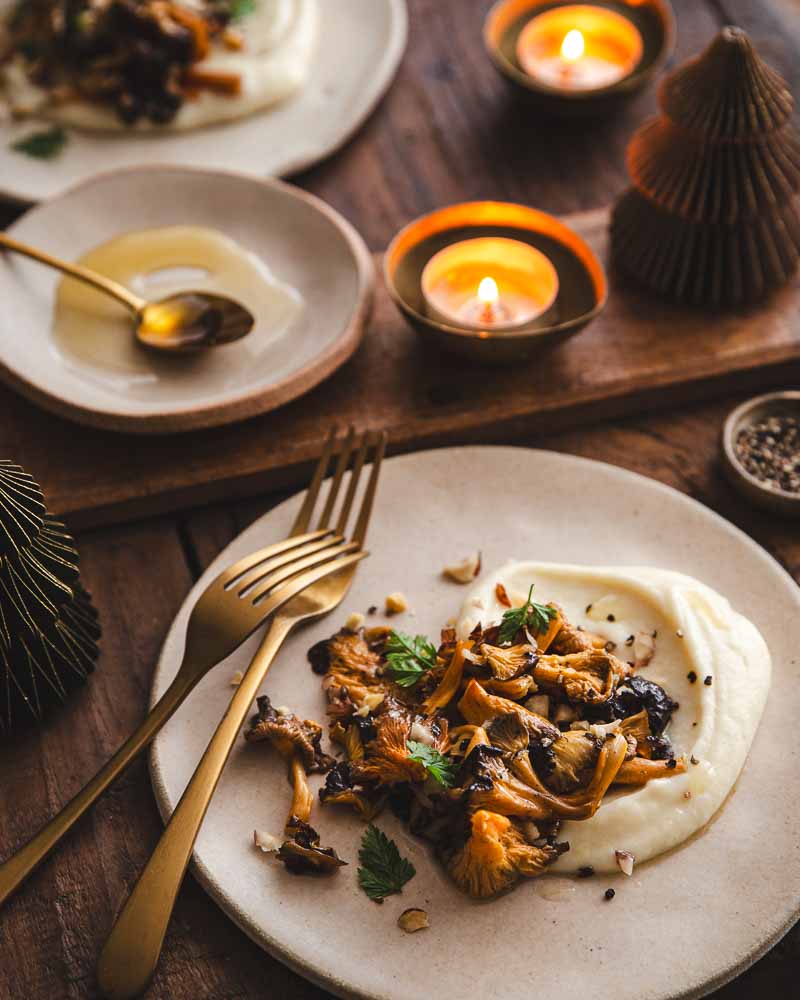 Une recette vegan pour les fêtes : poêlée de chanterelles aux noisettes concassées avec une crème de panais et de l'huile de noisette disposée dans une assiette en céramique avec deux fourchettes dorées l'une à côté de l'autre.  

En arrière plan, une coupelle en céramique avec de l'huile de noisette, une seconde assiette garnie, des photophores dorées, des sapins de noël en papier dans une ambiance de fin de journée