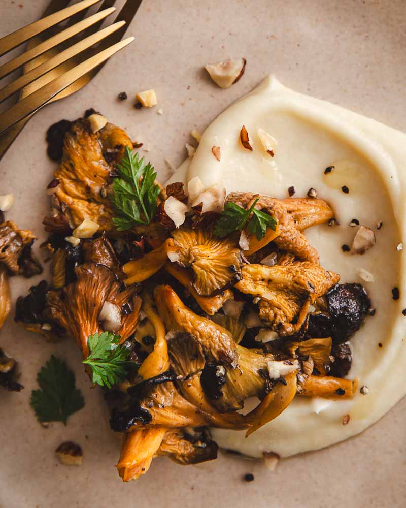 Gros plan sur une poêlée de champignons parsemée de noisettes concassées. Au fond de l'assiette, une crème de panais végétale. 