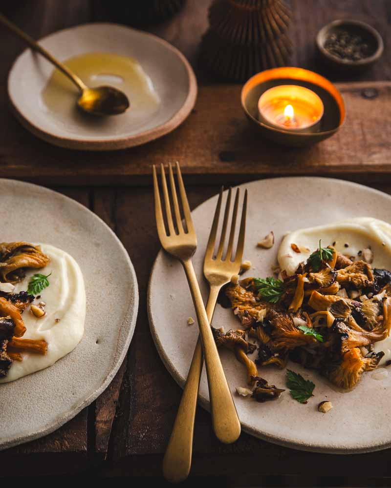 Deux assiettes en céramique côte à côte avec deux fourchettes dorées croisées garnies d'une purée de panais à la crème de soja et d'une poêlée de chanterelles, d'éclats de noisettes et d'huile de noisette avec du cerfeuil. 
