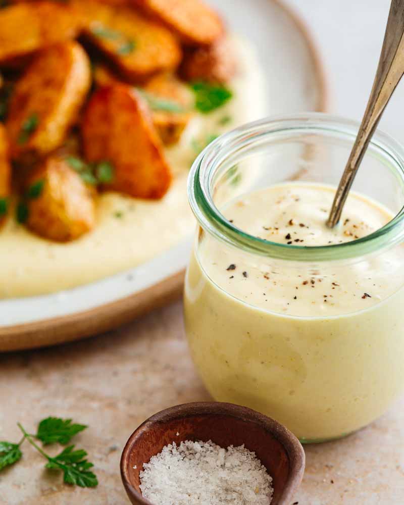 Pot en verre contenant la sauce végétale type mayo au tofu soyeux et moutarde forte avec des grains de poivre. En arrière plan, une assiette de pommes de terre rôties aux épices à l'Air Fryer