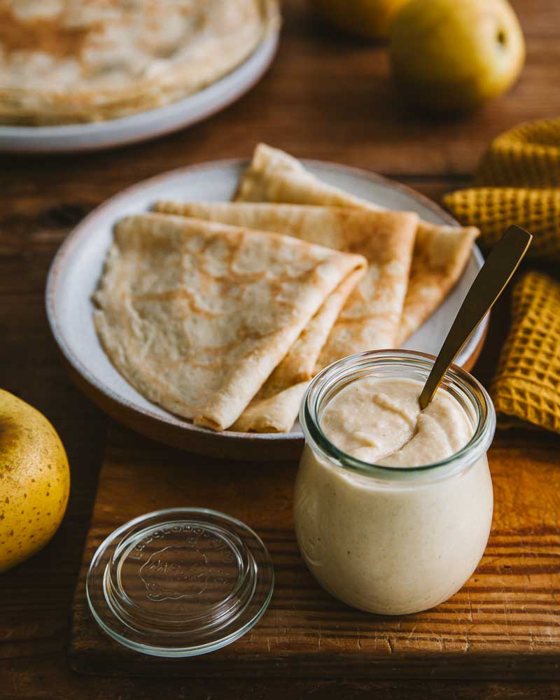 Pot de beurre de pomme végétal en premier plan, en arrière plan une assiette avec des crêpes pliées prêtes à être dégustées. 