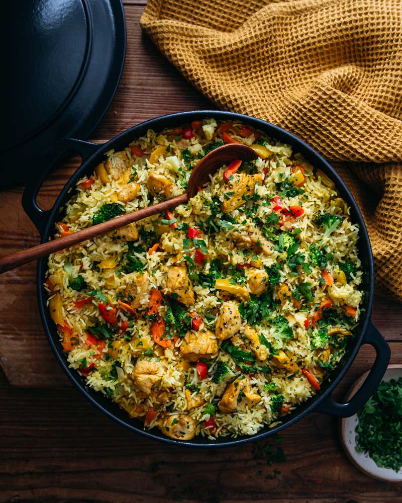 Vue de haut d'une cocotte en fonte noire contenant un plat complet composé de riz, de poivrons rouges et jaunes,  de brocolis et de carottes ainsi que de bouchées de blancs de poulet aux épices.  Un peu de persil est parsemé sur le plat et une cuillère en bois en posée dans la cocotte. Autour, le couvercle en fonte, un tissu gaufré couleur curry sur une table en bois. 