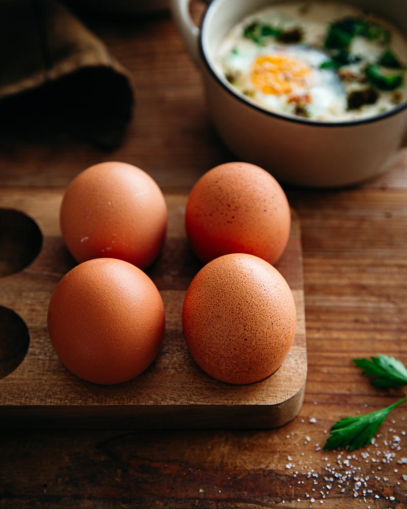 4 oeufs extra-frais posés sur un socle en bois pour les oeufs. En arrière plan, un ramequin d'oeuf cocotte aux brocolis et crème végétale au soja dans un ramequin beige en céramique. 