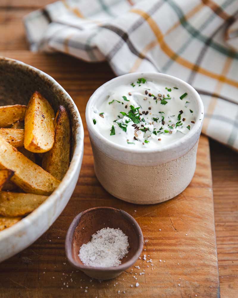 Pot en céramique beige et blanc contenant une sauce au yaourt à la grecque posé sur une planche. A côté un bol avec des potatoes au paprika cuites à l'Air Fryer. 