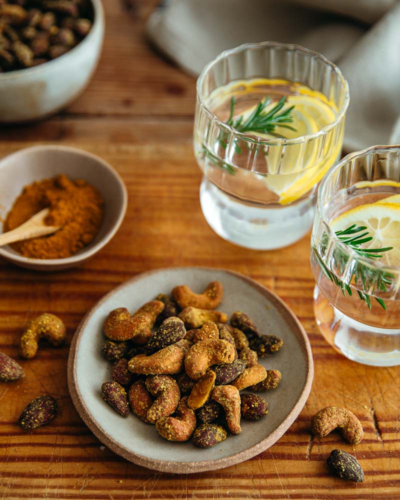 Coupelle en céramique remplie de noix de cajou et amandes au curry grillées à l'Air Fryer. A côté, coupelle de curry et verres avec rondelles de citron et romarin. 